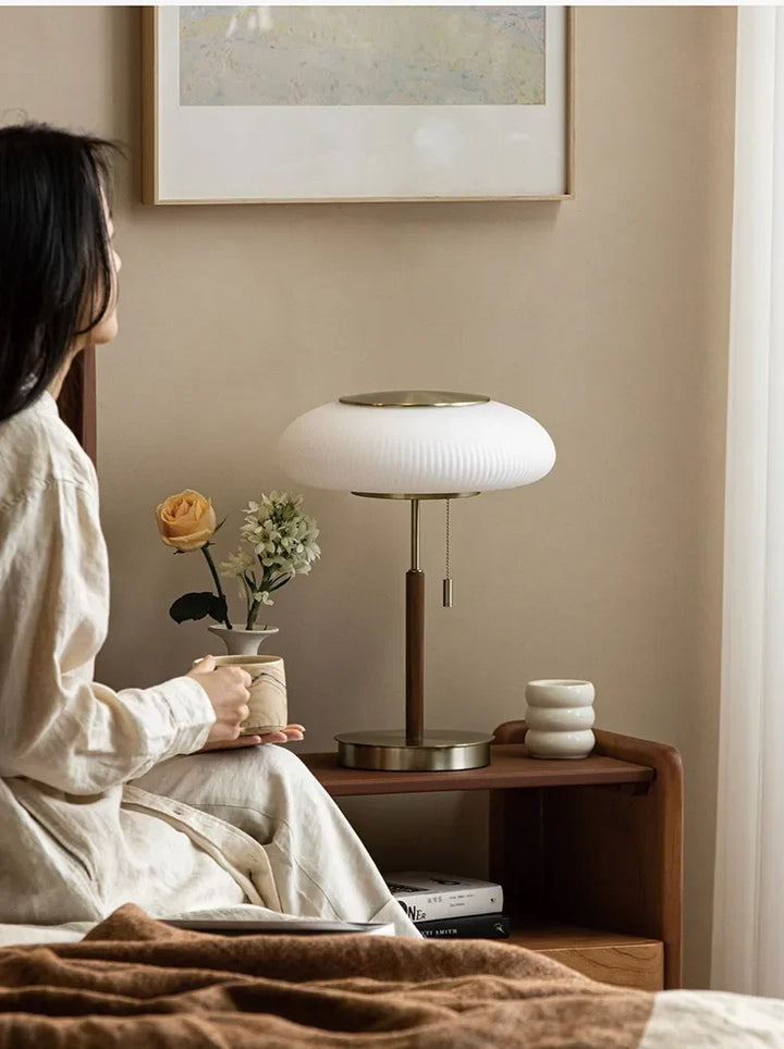 Woman sitting on a bed holding a mug with a lamp and flowers on a nightstand in the background.