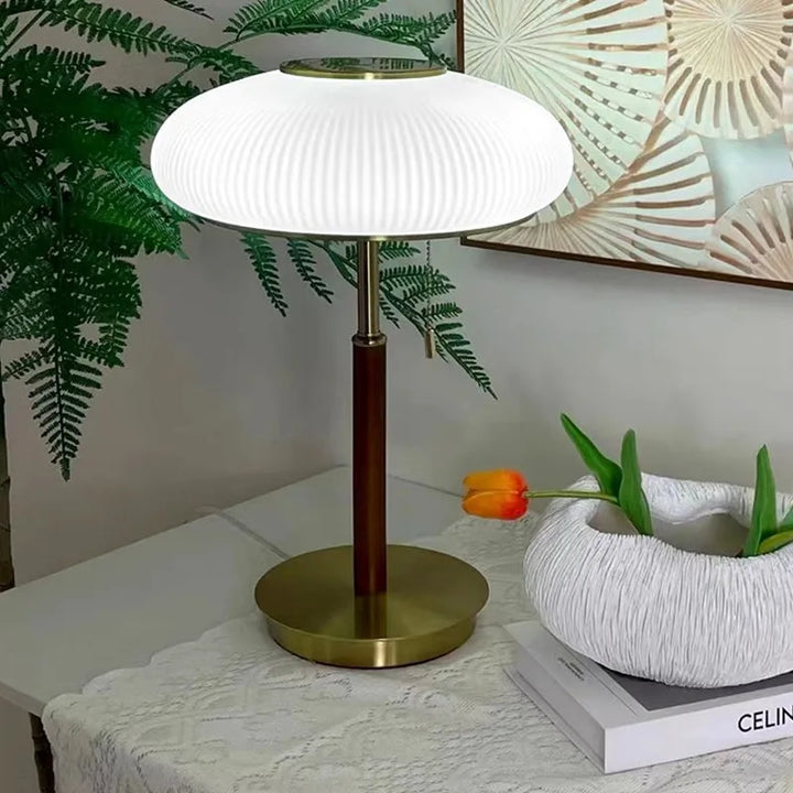 Table lamp with a white shade and gold base on a table with a plant and books in the background.