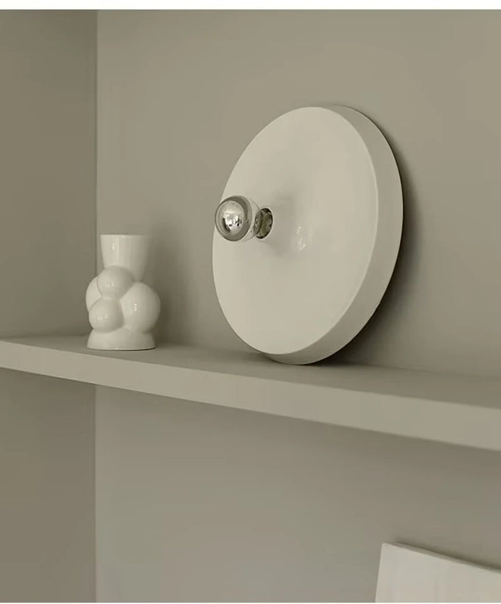 Round white wall-mounted light fixture on a shelf against a beige wall.