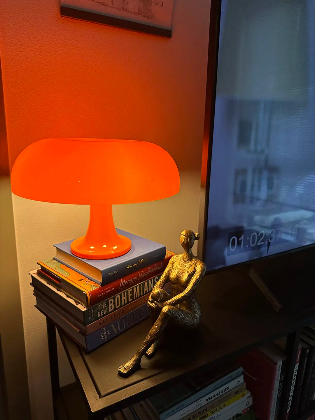 Orange lamp on books with a gold sculpture in front of a television displaying a video.