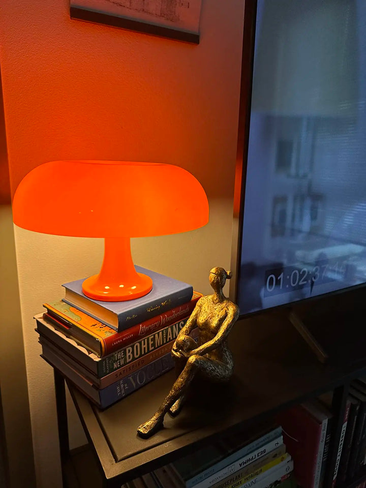 Orange lamp on books with a gold sculpture in front of a television displaying a video.