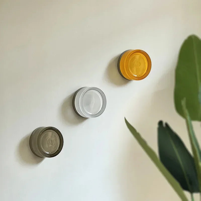 Three circular wall lights in different colors on a white wall with a plant in the corner.